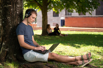 A blond young male student working on his laptop sitting under a tree in the park.