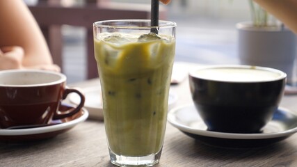 Stirring matcha ice tea latte with plastic straw. Two mugs of coffee on the background