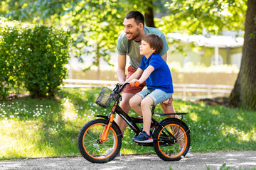 family, fatherhood and leisure concept - happy father teaching little son to ride bicycle at park
