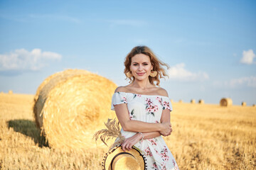 Naklejka na ściany i meble Young blond woman, wearing long white romantic dress, holding straw hat and dried grass bouquet, posing in front of wheat bale on field in summer. Female portrait on natural background.