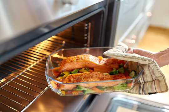 food cooking, culinary and people concept - young woman with potholder taking baking dish with salmon fish and vegetables out of oven at home kitchen