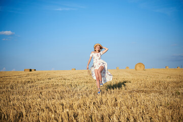 Young blond woman, wearing white romantic dress and straw hat and dried grass bouquet, jumping running on straw field in summer. Female portrait on natural background. Environmental protection.