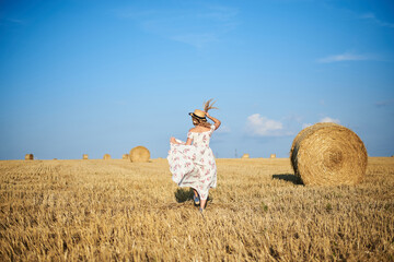 Naklejka na ściany i meble Young blond woman, wearing white romantic dress and straw hat and dried grass bouquet, jumping running on straw field in summer. Female portrait on natural background. Environmental protection.
