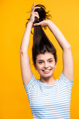 Fototapeta premium brunette woman holding hair above head and smiling isolated on yellow