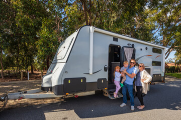 A Young Family proudly standing outside of their  caravan in front of a Park - obrazy, fototapety, plakaty