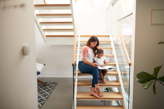 Hispanic Mother And Daughter Sitting On Staircase In Modern Home Reading Book Together