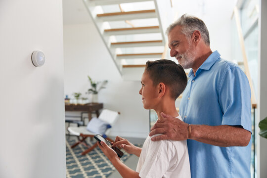 Grandfather With Grandson Adjusting Digital Central Heating Thermostat In Home