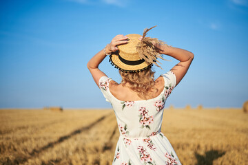 Young blond woman, wearing white romantic dress,with back to camera, holding straw hat and dried grass bouquet on head, walking on straw field in summer. Female portrait on natural background.