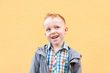 A red-haired boy laughs and looks to the side and up, dressed in a plaid shirt and a gray jacket, stands on the street against a yellow wall