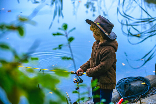 Little boy fishing in overalls from a dock on lake or pond. Child with a fishing  rod standing by the water. Young fisherman on a fishing trip, back view.  Stock Photo