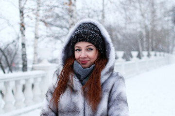 Young beautiful woman with red hair and blue eyes in gray faux fur coat with hood on background of winter snowy park.