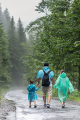 Beautiful mountain landscape. Father, son and daughter are walking on a wet road after a heavy rain in raincoats. The boy is holding his father's hand.