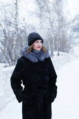 Fototapeta premium Young beautiful woman with short blond hair in black faux fur coat with gray fluffy hood and black knitted hat is walking in winter snowy park.