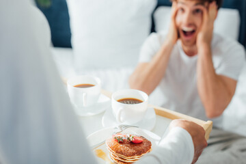 Selective focus of woman holding breakfast and coffee on tray near excited boyfriend in bedroom