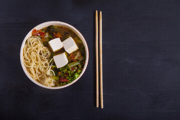 Fototapeta na wymiar Japanese soup with tofu, noodles, vegetables and herbs, bamboo chopsticks on a black background. Copy spaes.