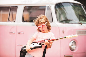 Child musician playing the guitar like a rockstar on pink background in neon light. Caucasian little boy learning to play guitar.