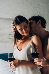 Selective focus of shirtless man kissing seductive woman with glass of wine in bedroom