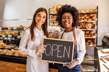 Happy businesswomen standing at bakery shop with open signboard. Close up of women holding sign now...