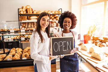 Portrait of two young entrepreneurs standing in the bakery shop with OPEN sign. Two cheerful small business owners smiling and looking at camera while standing with open sign board. - Powered by Adobe