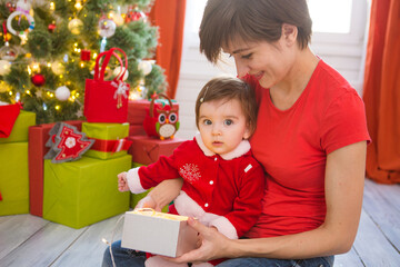 Young mother and her little daughter dressed as santa claus opening a magical Christmas gift by a Christmas tree in cozy living room in winter