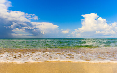 The turquoise sea washes the sandy beach. In the background there is a blue sky and a thundercloud.