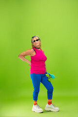 Fototapeta na wymiar Skater girl. Senior woman in ultra trendy attire isolated on bright green background. Looks stylish and fashionable, forever young. Caucasian mature woman in sunglasses, bright attire and sneakers.