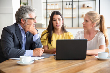 Young entrepreneurs and mature investor watching presentation and discussing project. Man and women meeting at co-working and using laptop. Medium shot. Partnership and investment concept