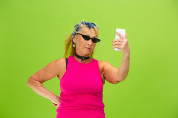 Taking selfie. Senior woman in ultra trendy attire isolated on bright green background. Looks stylish and fashionable, forever young. Caucasian mature woman in sunglasses, bright attire and sneakers.