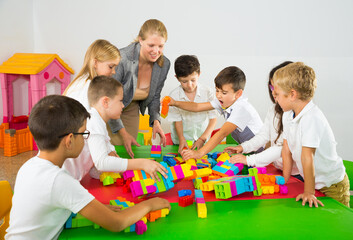 Fototapeta premium Happy kids and female teacher playing together with colorful toy building blocks in classroom at elementary school