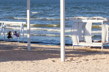 Children swing on swing by sea. Canopy with white curtains on sandy beach by sea.