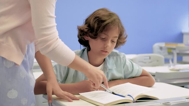 Boy reading textbook in class. A girl helps him and explains the exercises to do. Medium shot. Child sitting at his desk.