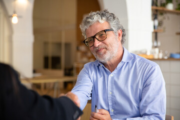 Positive friendly businessman in glasses shaking hands with partner at meeting. Medium shot....