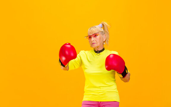 Boxing in gloves. Senior woman in ultra trendy attire isolated on bright orange background. Looks stylish and fashionable, forever young. Caucasian model in sunglasses, bright attire and sneakers