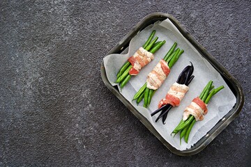A baked appetizer, a bundle of green beans wrapped in bacon in a metal baking dish against a dark concrete background. Hot appetizer recipes.