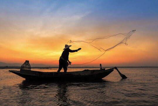 Silhouette of asian fisherman on wooden boat in action throwing a net for catching freshwater fish in nature river in the early morning before sunrise