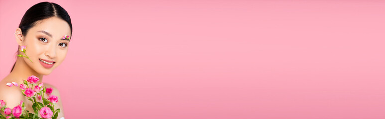Fototapeta premium horizontal concept of asian woman with buds and petals on face and shoulder, holding tiny roses isolated on pink