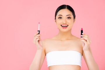 excited asian woman holding lip gloss and lipstick while looking at camera isolated on pink