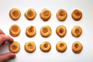 Fifteen handmade cookies with apricot jam arranged in even rows on white background. Man hand takes one from side.