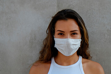 Portrait of young woman of Arabic ethnicity wearing disposable face mask as barrier to protect against contact with infectious materials standing near concrete wall. Close up, copy space, background.