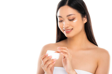 brunette asian woman touching cosmetic cream while applying it on face isolated on white