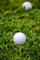 Close-up of golf cue ball on tee, with one ball out of focus, on grass field, in vertical with copy space