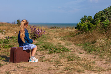 Naklejka na ściany i meble Little blonde girl in denim jacket, blue dress with vintage suitcase and flowers bouquet off-road with sea landscape. Stylish hitchhiker child with long hair on countryside trip. Kid walking outdoors.