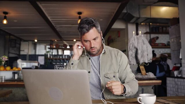 Handsome business man wearing earphones listening to video call on laptop while sitting with coffee at trendy cafe. - Powered by Adobe