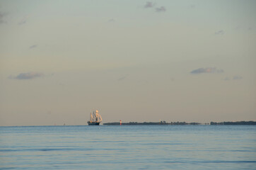 Old sail ship with full sails pass a distant light house, out in the horizon.