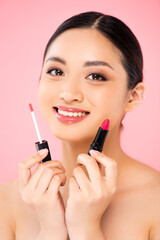 young asian woman looking at camera while holding lip gloss and lipstick isolated on pink