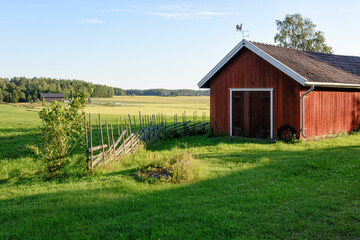 Traditional Finnish red wooden barn with roundpole fence and the weather vane on the roof. Old idyllic farm in the countryside. © Finmiki