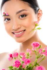 Fototapeta premium young asian woman with floral decor on face looking at camera near pink roses isolated on white
