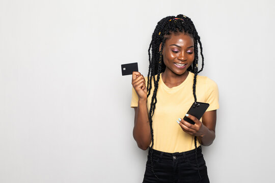 Picture of smiling young african woman using phone and holding debit card standing isolated over white background.