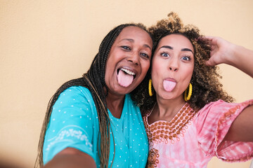 Beautiful african mother and adult daughter stick their tongue out while they take a selfie -...