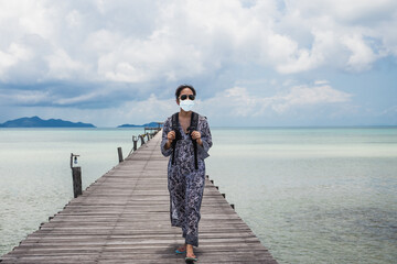 Tourist woman with protective mask walking on a wooden bridge with ocean view.